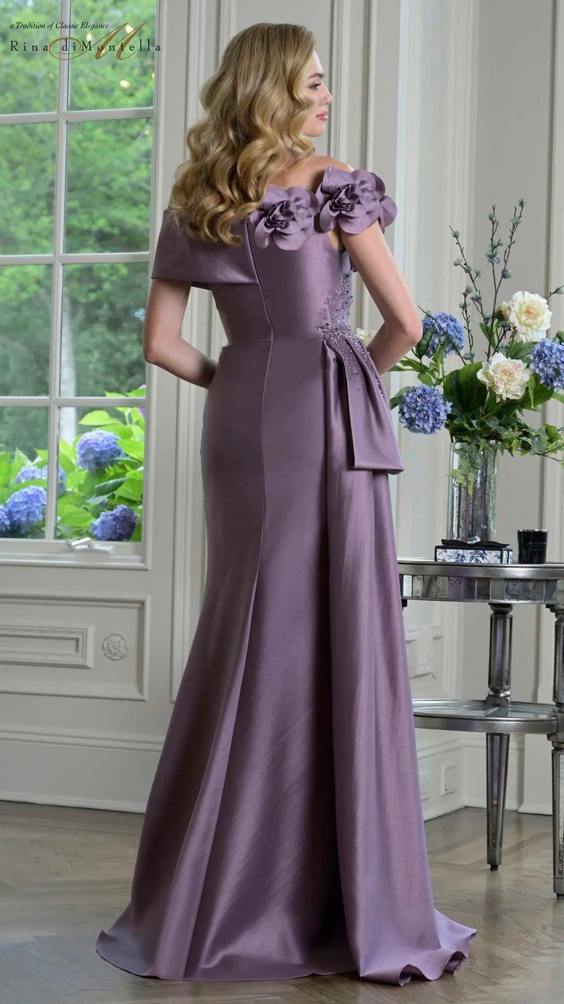 A woman wears the Rina Di Montella RD3407 Long Formal Dress, standing indoors by a window with flowers on a table in the background.